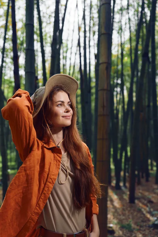Woman in forest wearing straw hat and orange jacket, smiling portrait among tall trees and autumn foliage with sunlight. Natural outdoor scene conveying peaceful nature and style.