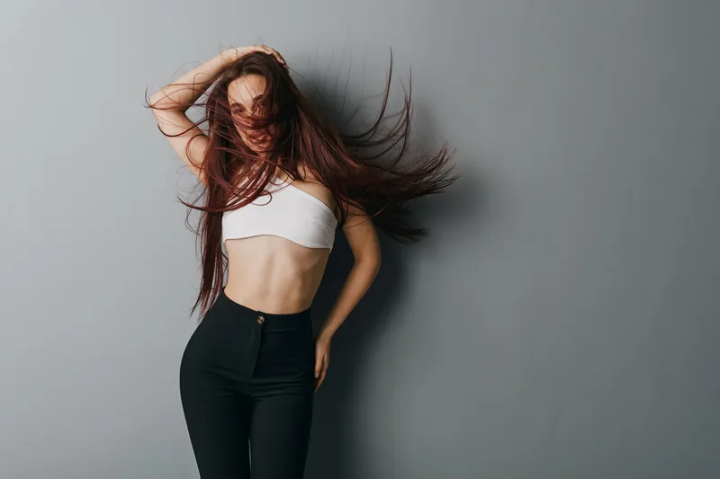 Woman with long hair wearing white crop top and black pants posing against gray background. Movement and fashion concept with casual style and confident female model in studio.