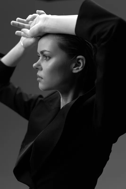 Black and white portrait of a serious woman in a blazer with raised arms, showing strong emotion and dramatic lighting.