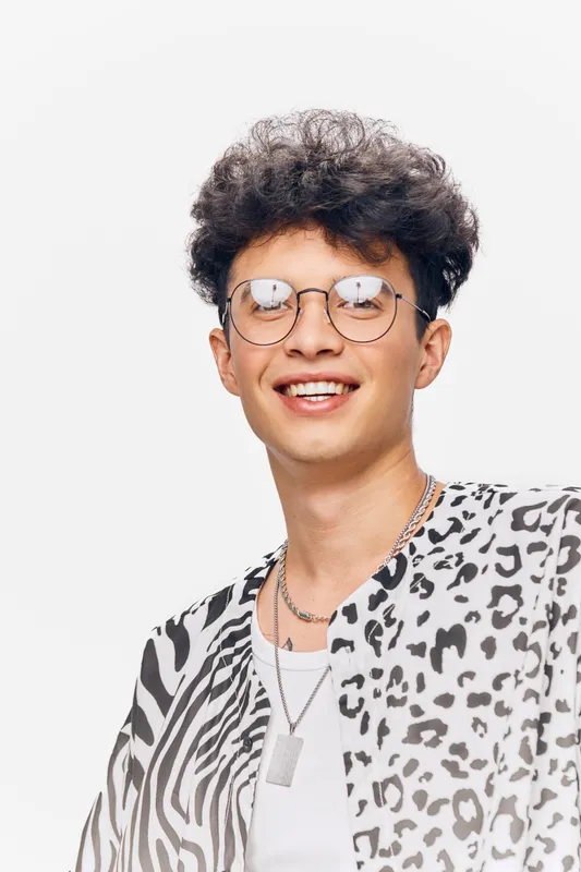 young man smiling with stylish curly hair wearing glasses and black and white patterned shirt, positive expression on bright white background