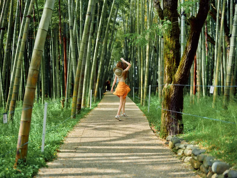 Woman in orange dress and straw hat walking along a bamboo path in a lush forest, summer travel scene with sunlight, greenery and a serene walkway for tourism and nature exploration.