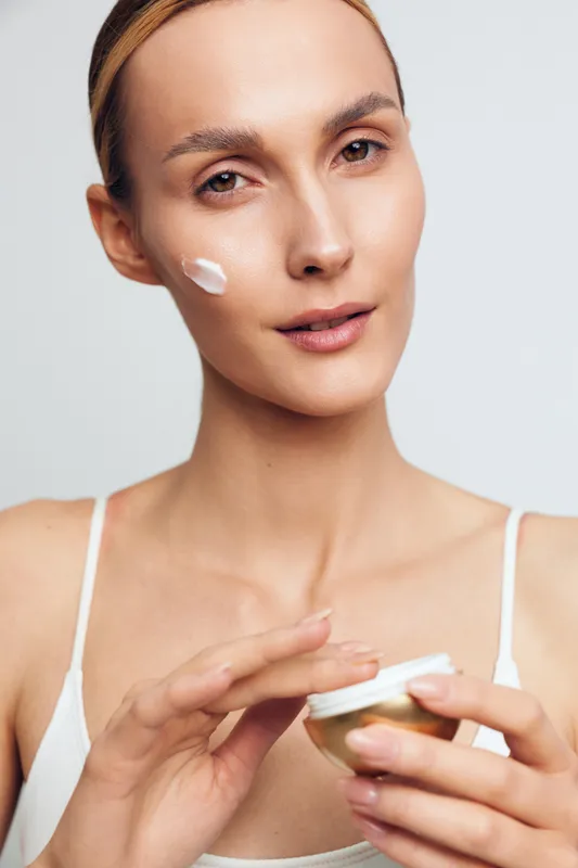 Woman skincare face cream spa close up beauty self care, holding jar, applying moisturizing cream to cheek, soft studio light, smooth skin, calm gaze, clean background