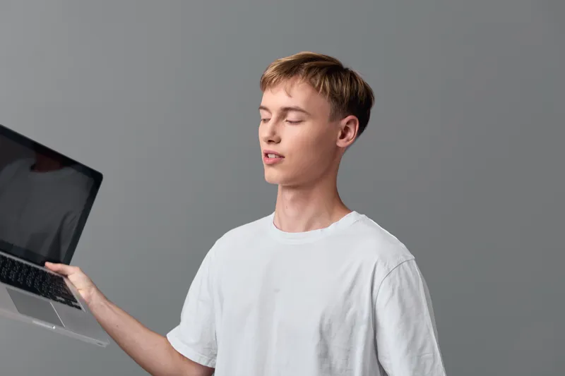 Laptop in hand, man in a white tee stands before a neutral gray backdrop, showcasing modern technology, work, and everyday productivity in a minimal studio setting