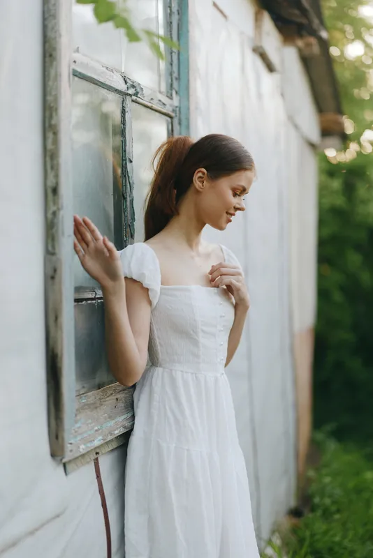 Young woman in a white dress standing by a rustic window, exuding a serene and contemplative mood amidst a lush green background