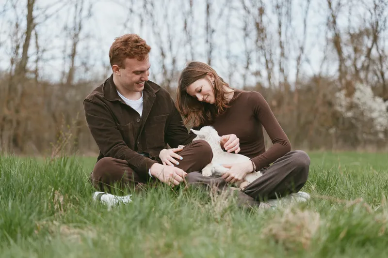Couple outdoors in a grassy field with a friendly goat kid, sharing a tender moment, calm and bright, showcasing a lifestyle moment with animals in nature, peaceful and intimate scene
