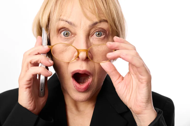 Senior woman holding glasses near eyes while talking on a phone, surprised expression in studio portrait closeup on a white background, communication moment, adult hands face