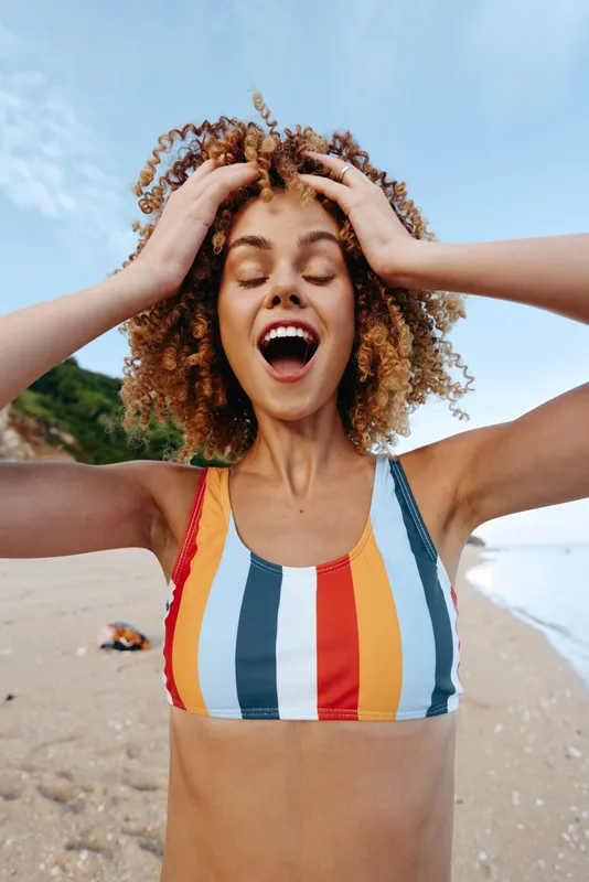 Joyful woman on a sunny beach wearing a striped bikini, hands in her hair, laughing with eyes closed, wind-swept hair, blue sky, relaxed summer moment with natural colors, soft light