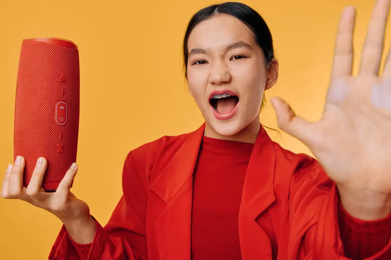 Young woman in red jacket holding red portable speaker with hand raised, expressing excitement and energy against orange background for music and technology concept.