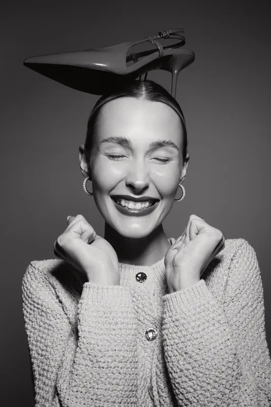 Happy white woman with fair skin smiles with eyes closed wearing a knitted sweater and high heel shoe on her head, studio portrait in black and white.