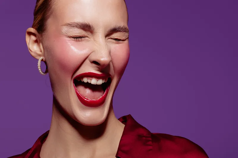 Close-up of happy woman with red lipstick laughing with eyes closed on purple background. Concept of joy, beauty, makeup, and positive emotions in studio portrait.
