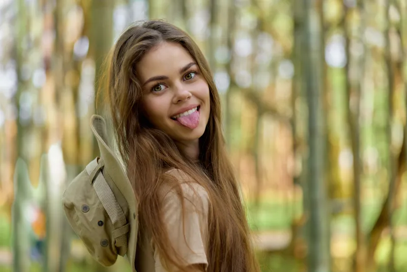 Girl portrait playful expression sticking tongue in forest with long hair and hat, casual outfit and natural light capturing youthful fun, candid smile and carefree outdoor mood.