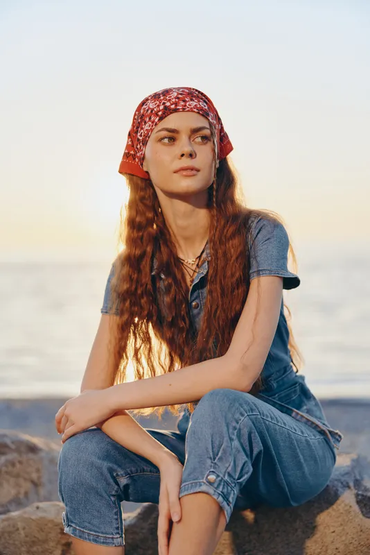 young woman in denim outfit wearing red bandana sitting on rock by the sea during golden hour with calm expression and natural light