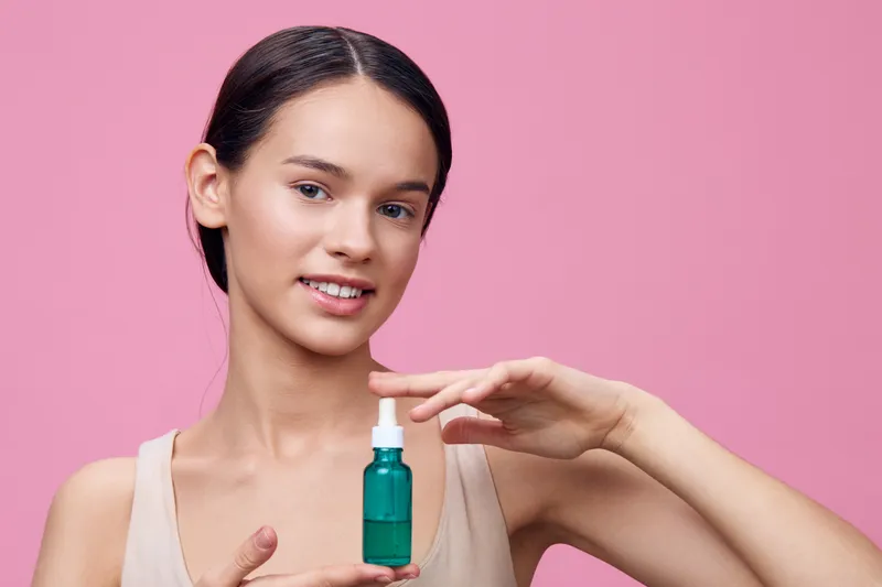 A young woman holding a green serum bottle against a pink background, showcasing healthy skin care and beauty, promoting self care and wellness
