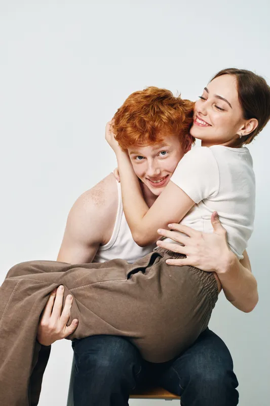Young couple embracing with happiness, smiling woman holding redhead man in casual clothes, joyful emotion and affection in studio portrait with white background and positive mood.