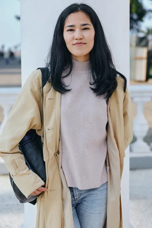 Woman outdoors in a beige coat, soft sweater, and jeans, carrying a shoulder bag, relaxed pose in urban light, showcasing casual street fashion and confident mood