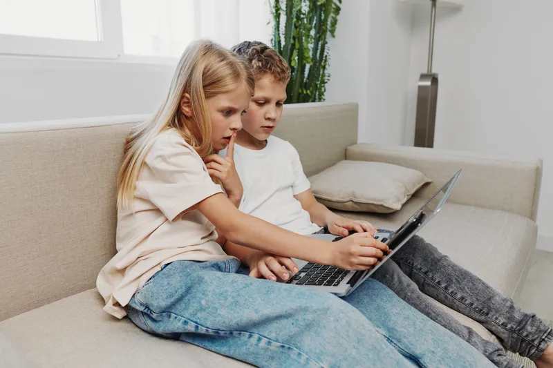Two curious children sitting on a sofa use a laptop together in a bright modern room, focused on the screen. Concept of learning, technology, and childhood curiosity.