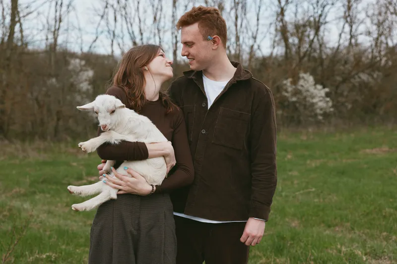 Young couple stands in a grassy field holding a white baby goat while looking at each other with gentle smiles during a calm spring day outdoors.