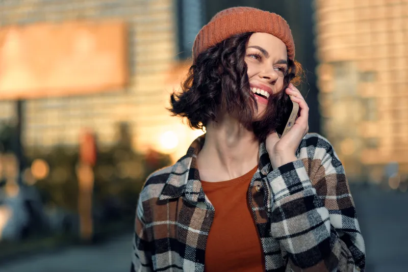 woman phone smile city beanie jacket, young woman laughing on mobile in urban golden hour glow, candid authenticity and emotional storytelling for mindful living and modern lifestyle