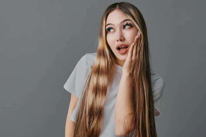 Young woman with long hair wearing white t shirt looking surprised with hand on cheek against gray background. Beauty, emotion and casual style for portrait and expression.