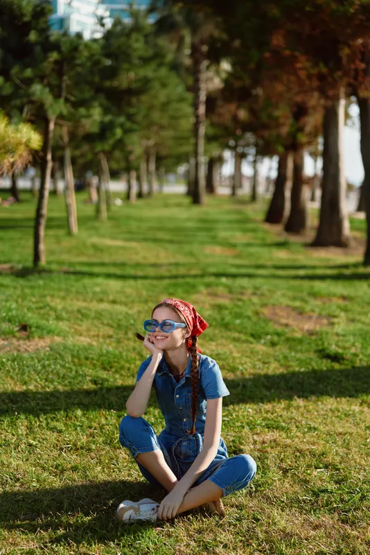 lifestyle woman in modern boho-western denim street style sitting on grass outdoors with film color effect under bright sunlight in urban park area