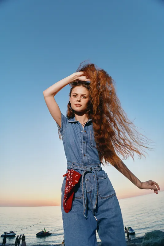 Lifestyle woman with modern boho-western denim street style, standing by the sea with film color tones and flowing red hair in natural sunlight at sunset.