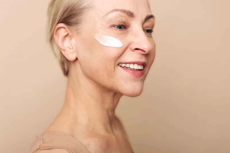 Smiling mature woman with light skin applies white face cream on cheek. Close-up on beige background. Concept of skincare, antiaging, beauty routine, and natural wellness for older adults.