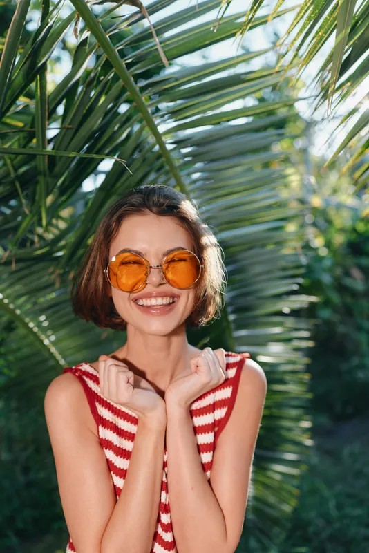 A cheerful woman outdoors wears round orange sunglasses and a striped sleeveless top. She smiles brightly, enjoying warm sunshine amid lush palm leaves, exuding a vibrant summer vibe and relaxed