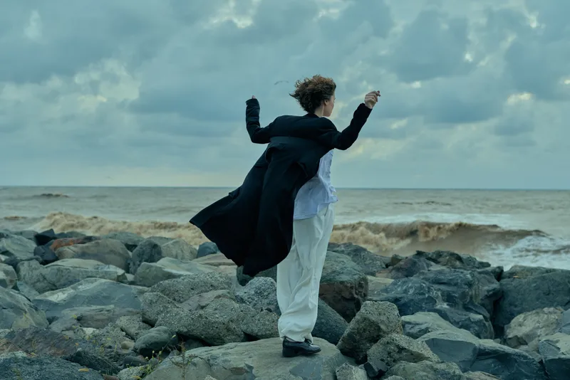 Couple embraces on rocky coast by the ocean, wind blowing coats as waves crash, dramatic moment of connection and freedom against a gray sky.