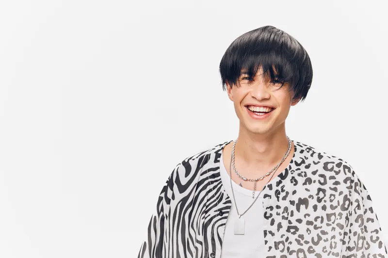 Smiling man with black hair wearing a stylish animal print shirt and layered silver necklaces, expressing happiness and modern fashion in a clean white background.