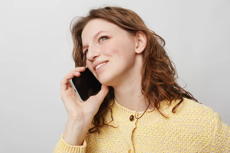 Smiling woman with curly hair wearing yellow sweater talks on smartphone against white background. Concept of communication, happiness, casual lifestyle, and positive emotions.