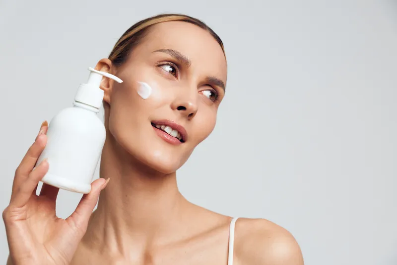 Young woman with a clean complexion applies white face cream from a pump bottle on her cheek, smiling softly against a neutral background