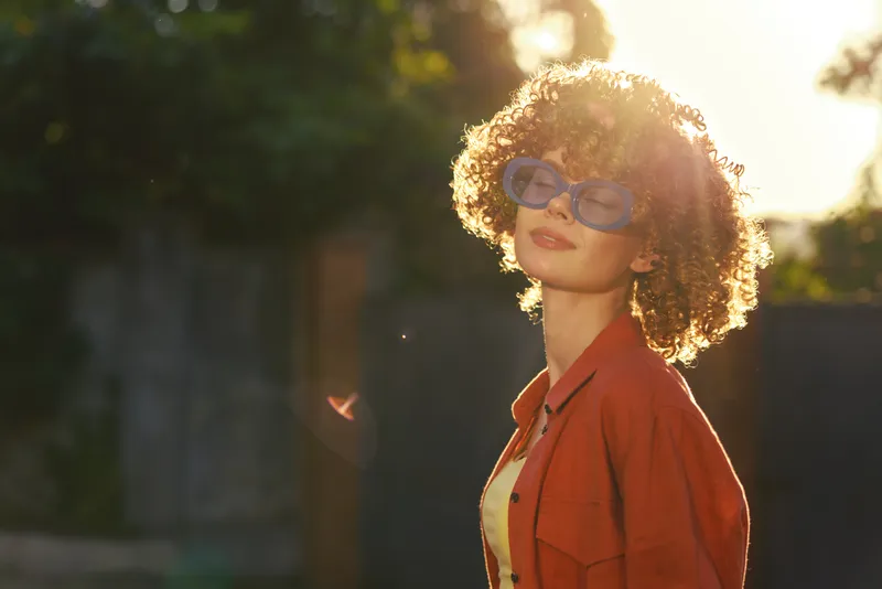 Smiling woman with curly hair wearing sunglasses outdoors in warm sunlight. Casual fashion, solar glow, carefree lifestyle, summer mood, soft backlight