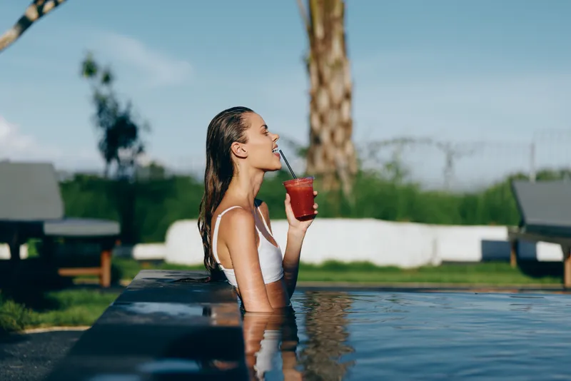 Smiling woman in a white swimsuit drinks a red beverage by the pool, under sunny outdoor skies, capturing a carefree lifestyle and refreshing summer mood.