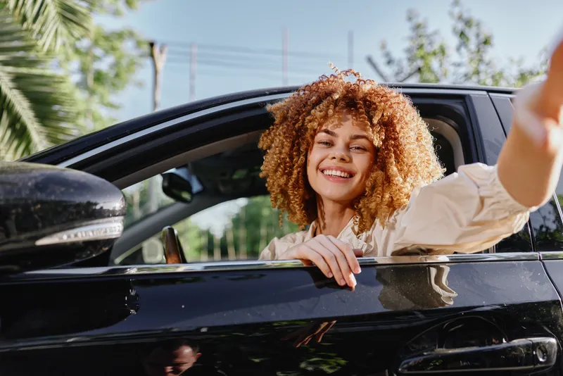 Smiling woman with curly hair sitting in black car, enjoying sunny day outdoors with relaxed and happy expression, modern lifestyle and freedom concept.