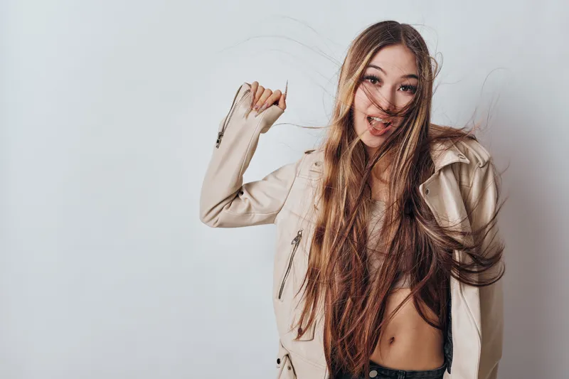 Young woman with long brown hair wearing light jacket posing cheerfully with hair flying against white background in studio for fashion portrait.