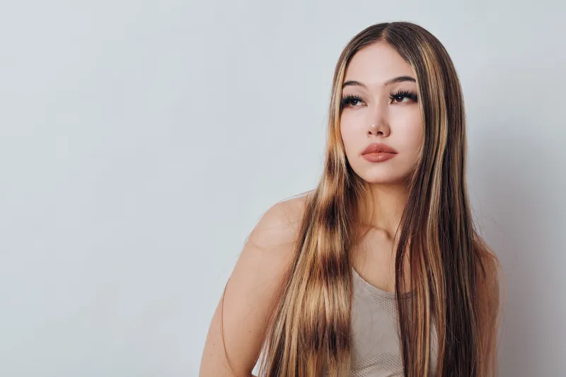 Woman with long hair and makeup posing against light background, portrait of young female model with straight hair and natural expression in studio photography.
