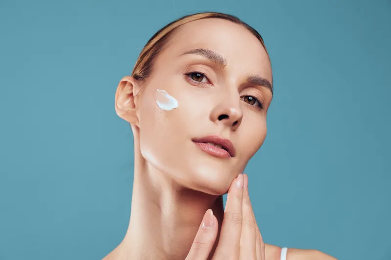 Woman applying white face cream on cheek with fingers. Close-up portrait on blue background. Concept of skincare, beauty, wellness, and self-care with soft natural lighting.
