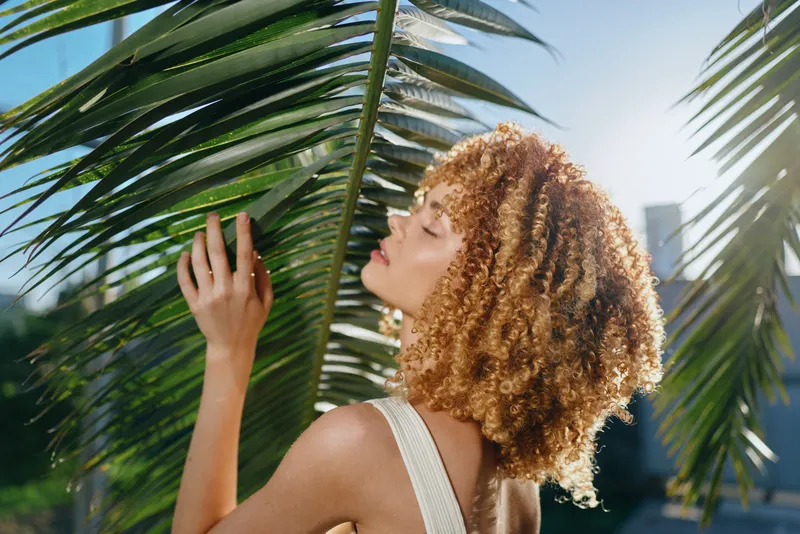 young woman with curly hair enjoying sunlight under green palm leaves in summer atmosphere with blue sky and soft natural light