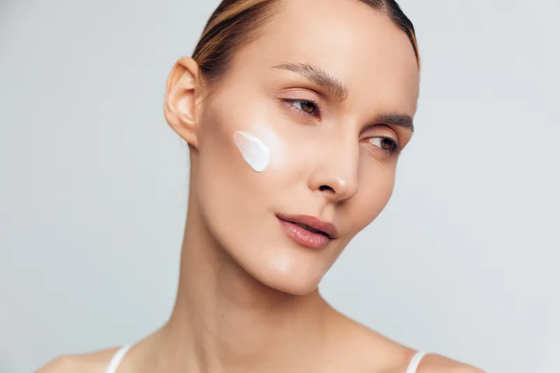Young woman with brunette hair applies white face cream on her cheek while looking away against a plain light background, expressing calmness and self-care.