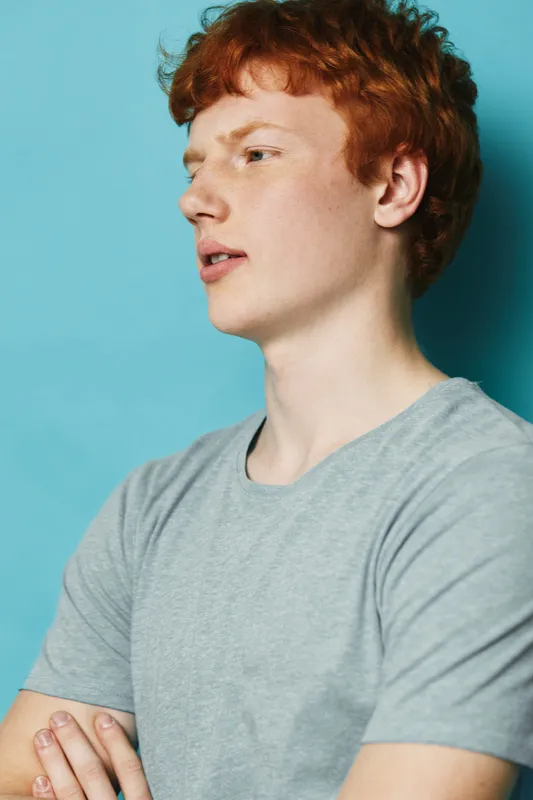 Young man with red hair and freckles wearing gray t shirt with crossed arms looking aside against blue background in studio. Casual style portrait of confident male person.