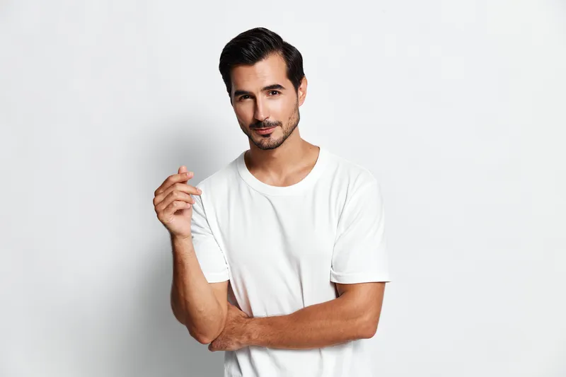 Handsome man with charming smile wearing a white t-shirt poses confidently while looking at the camera against a minimalist light grey background. Portrait concept.