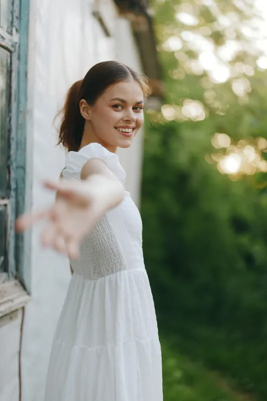 A young woman in a white dress smiling, reaching out with her hand amid a lush green background, conveying joy and warmth in a serene summer setting