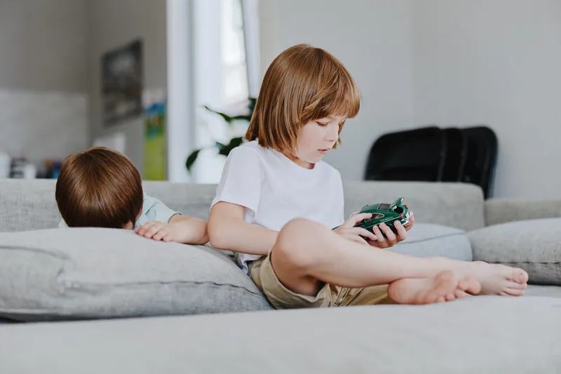 Engaging boy playing with a toy car while his younger sibling rests on the couch, surrounded by a cozy living room atmosphere filled with natural light.