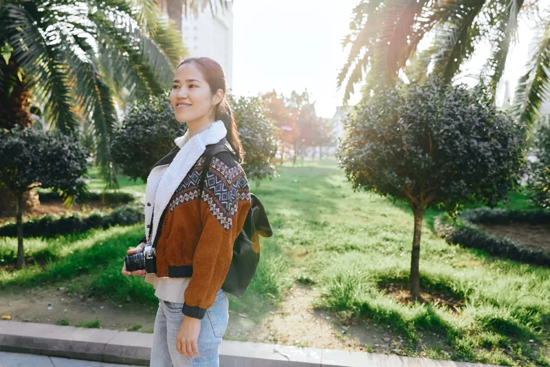 Woman with camera outdoors in a sunlit park, wearing casual fashion and a backpack, enjoying outdoor photography and a peaceful afternoon amidst greenery
