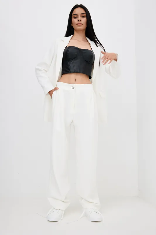 Woman in white pants suit and black corset top posing confidently against white background, showcasing modern fashion style with casual sneakers.