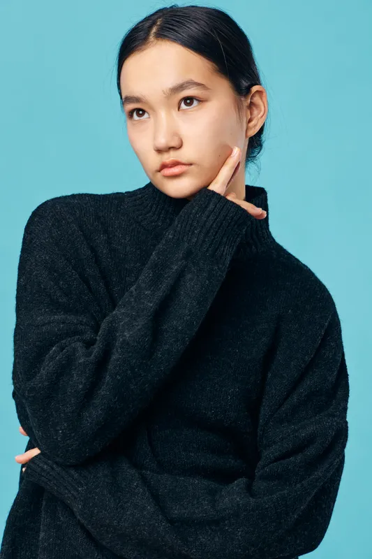 Young woman with black sweater posing on blue background, looking thoughtful with hand near face. Portrait of female model with natural makeup and dark hair pulled back in studio lighting.