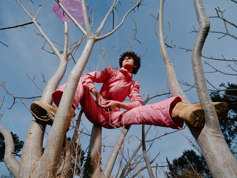 Person in a red outfit sits among leafless tree branches, arms spread for balance, high above ground. Outdoor nature shot under a bright blue sky with a bold adventurous mood.
