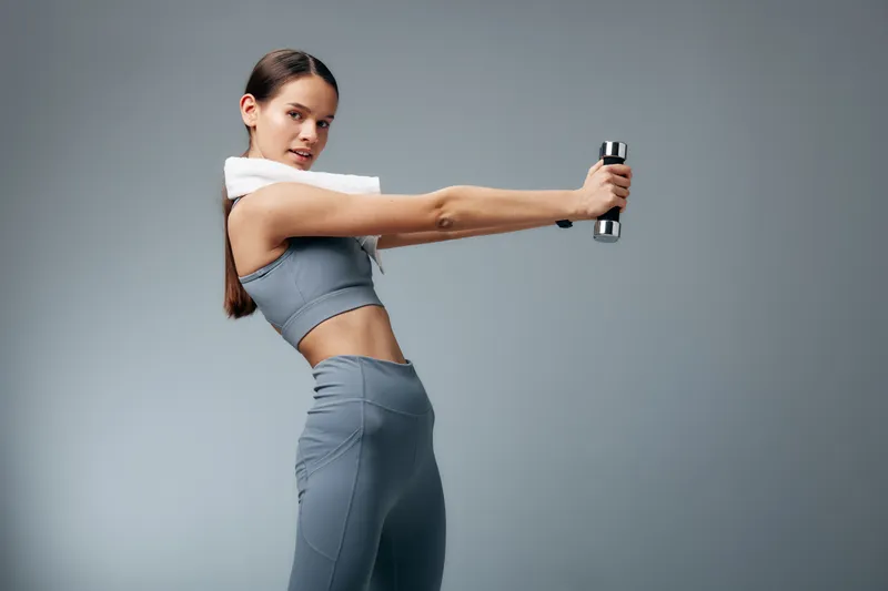 Athletic woman in a gray sports outfit demonstrates a dumbbell workout with a towel over her shoulder, facing forward in a minimalist studio for a dynamic fitness training scene.