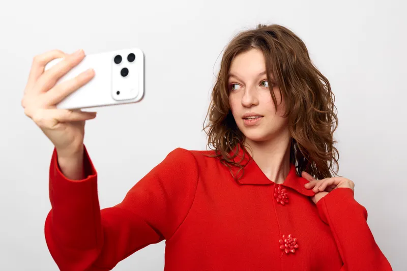 Young woman in red sweater takes selfie with smartphone on white background. Casual style, natural expression, and modern technology concept for social media and communication.