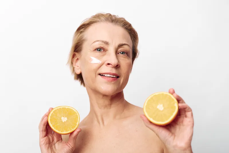 Mature woman with short hair holds two orange halves near face with cream applied on cheek, smiling against white background. Concept of skincare, natural beauty, wellness, and vitamin c care.